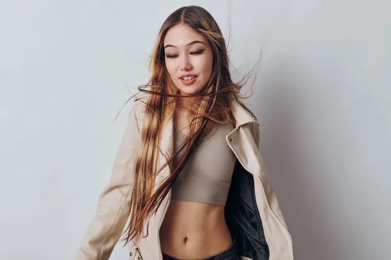 Young woman with long hair wearing beige jacket and crop top posing with eyes closed on white background for fashion and beauty portrait.