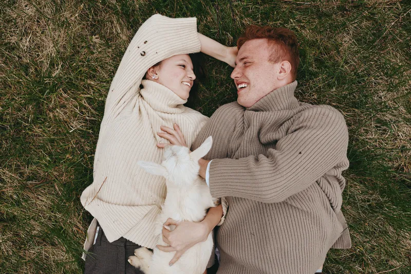 A joyful couple lying on a grassy field, smiling at a playful goat kid between them. A cozy outdoor lifestyle moment highlighting affection, simplicity, and bonding with animals in a natural setting.
