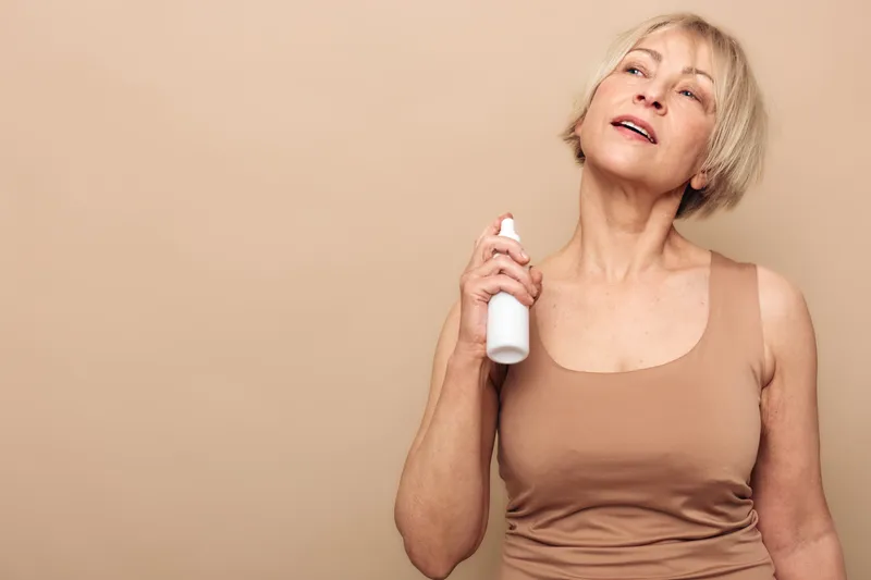Mature woman with short blonde hair sprays facial mist on her neck. Beige background and casual tank top. Concept of skincare, hydration, and self-care for aging skin.