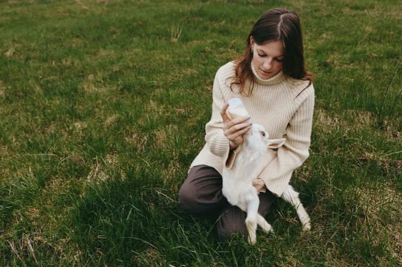 A woman sits on grass in a quiet field, bottle feeding a white goat kid, sharing a gentle intimate moment with a small animal, capturing a warm lifestyle scene amid nature and open space