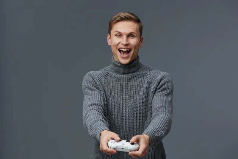 Happy young man in gray sweater playing video game with controller, smiling and showing excitement, indoor studio shot on plain gray background, lifestyle entertainment fun concept