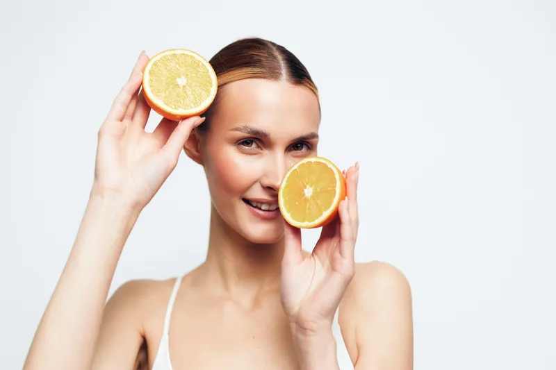 A young woman with brunette hair holds two orange halves up to her face, creating a playful, fresh moment in a bright, minimal studio with soft lighting and a clean backdrop.