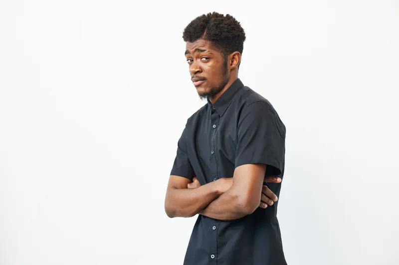 Black man with crossed arms, showcasing a confident demeanor. His stylish black shirt complements the minimalist background, enhancing the focus on his expression.