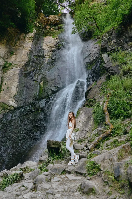 woman near waterfall standing on rocky trail surrounded by green forest and moss-covered stones, casual outfit, outdoor adventure and nature exploration.