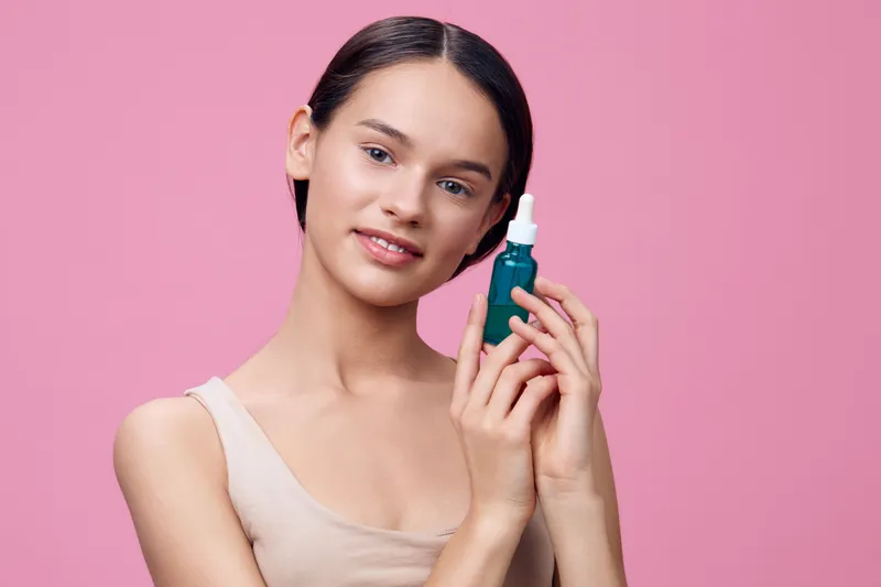 Young woman holding a blue serum bottle, smiling against a pink background, representing beauty and skincare Perfect for wellness and personal care concepts