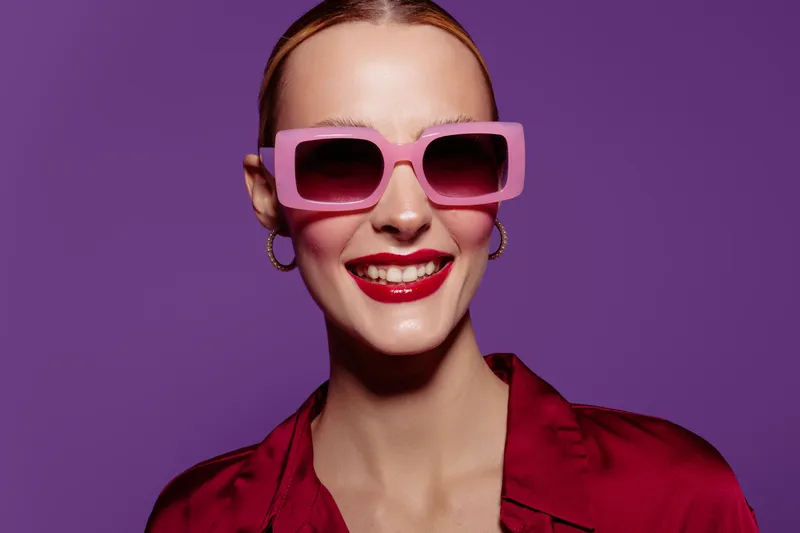 Smiling woman with red lipstick wearing pink sunglasses and red blouse poses against purple background. Concept of fashion, style, beauty, and confident female portrait.