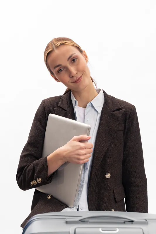 Young businesswoman with blonde hair holds laptop and stands near suitcase on white background. Concept of travel, work, and professional lifestyle with confident expression.
