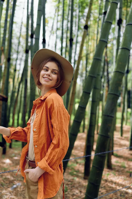 Woman in a bamboo forest smiling in a straw hat and orange shirt, portrait of a traveler enjoying nature. Casual outfit with hand in pocket, serene outdoor scene among tall green trees.