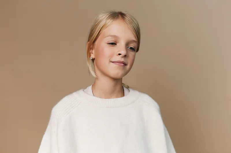 Portrait of a young girl with light blonde hair wearing a cozy white sweater, standing against a warm beige studio backdrop with a gentle, calm expression.