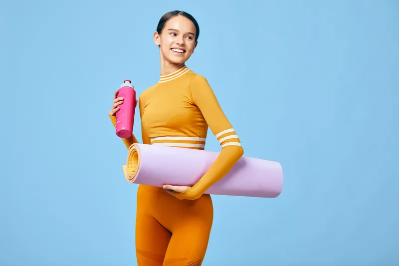 Athletic woman in orange sportwear poses with a pink water bottle and purple yoga mat against a bright blue studio backdrop, showcasing a dynamic fitness lifestyle.