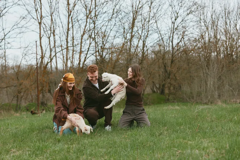Three young adults enjoy a playful moment outdoors with two baby goats in early spring, surrounded by bare trees and fresh green grass under a cloudy sky.