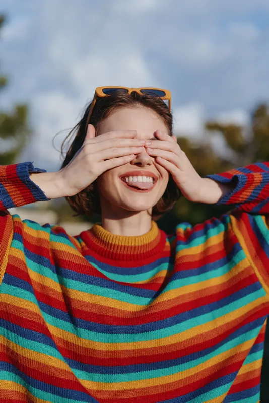 Woman smiling and covering eyes with hands in a colorful rainbow sweater, sunglasses on her head, playful outdoor portrait conveying joy, carefree mood and casual fashion.