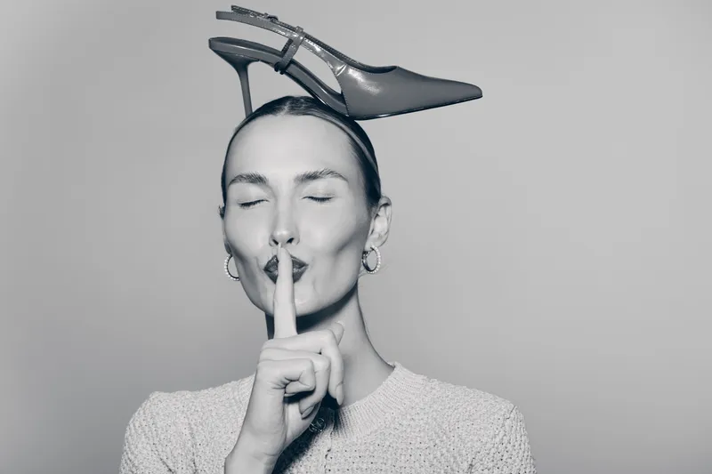 Portrait of a light skinned white woman with a shoe on her head, making a shushing gesture with closed eyes.