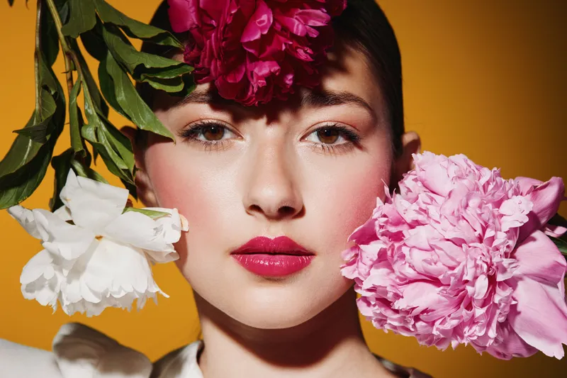 Bold beauty portrait of a young woman with bright pink lipstick and dewy skin surrounded by colorful blooming flowers on a yellow background. Creative fashion beauty concept.