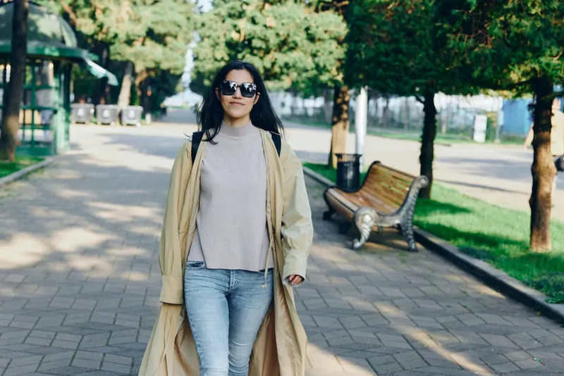 Woman in a long beige coat and light sweater walks through a sunny park along a paved path, showcasing casual fashion, chic style, and a relaxed outdoor urban lifestyle