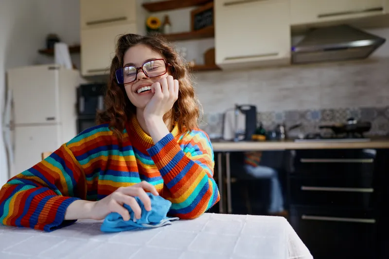 Cheerful woman indoors at a kitchen table, smiling while wiping with a cloth, colorful striped sweater, warm natural light, relaxed home moment, casual lifestyle, cozy ambiance indoors.