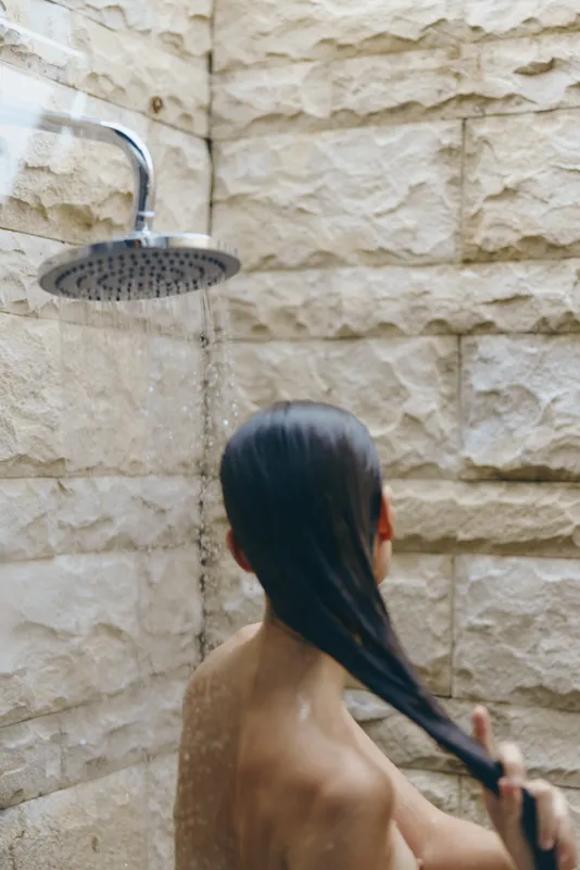 woman washing hair under outdoor shower with beige stone wall background, natural light, wet long dark hair, self-care moment, wellness concept
