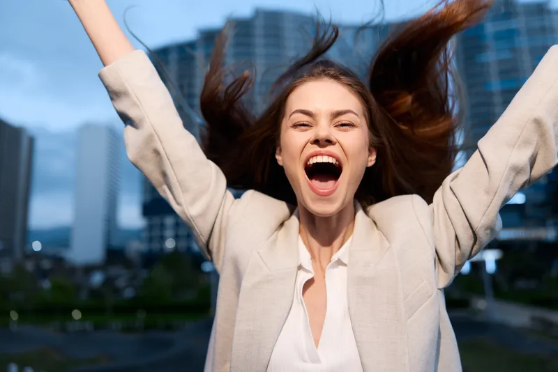 Excited woman in beige jacket celebrating success in urban business district with joyful expression and dynamic hair movement during daytime