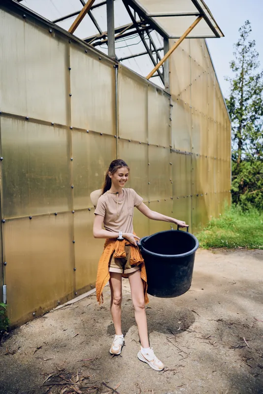 woman, girl and bucket near greenhouse gardening scene outdoors, casual young woman smiling while carrying a black tub for planting and watering in a backyard garden.