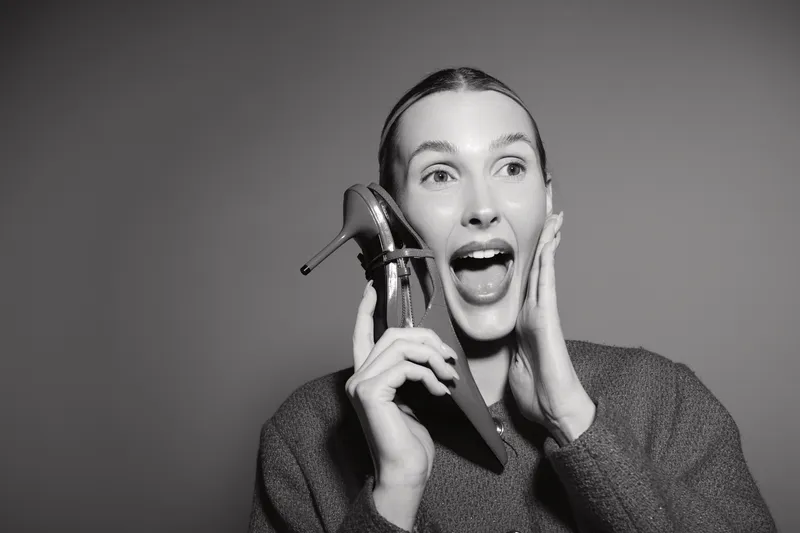 Young white woman with light skin and brown hair holds high heel shoe near face, expressing surprise and joy in studio on grey background. Concept for fashion and emotions.