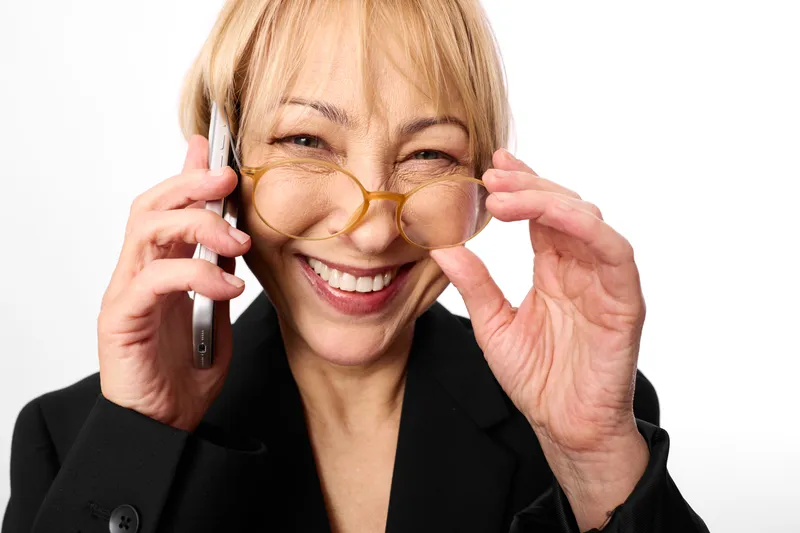 Woman, glasses, phone, call, smile, business, portrait indoors, cheerful laughter, warm studio lighting, looking at camera, relaxed hands, modern communication, office background