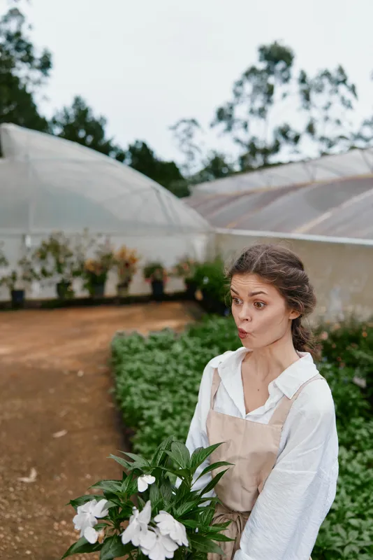 Young woman in a greenhouse with flowers, expressing surprise and joy, wearing a white shirt and apron, featuring a natural green background