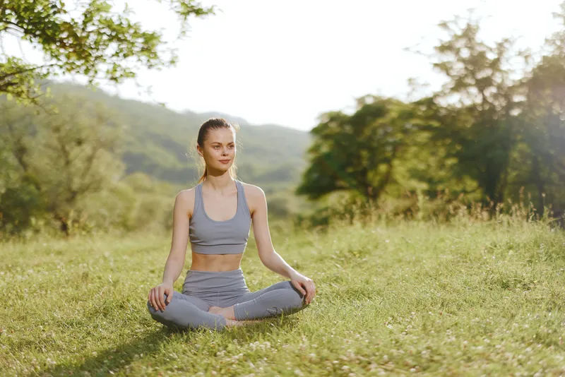 Female model practicing yoga in nature, wearing gray sportswear tranquil expression with green background promotes mindfulness and wellness through outdoor activities