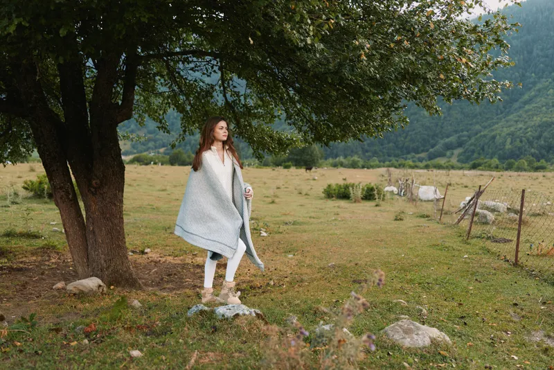 Woman lifestyle scene in rural meadow, standing near a tree, wearing a light shawl, serene pose, daytime natural landscape, open field and distant hills in bright sun