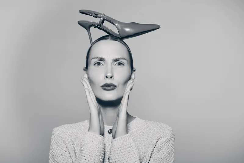 Portrait of a thin Caucasian woman with pale skin wearing a high heel on her head, posing with hands on face against plain background in black and white.