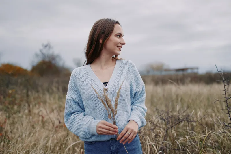 A candid portrait of a young woman in a windswept field, wearing a cozy knitted sweater and holding dry grass, conveying an authentic, realistic moment of calm and genuine emotion. (160 characters)