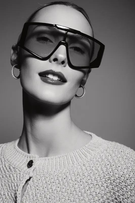Stylish white woman with fair skin wearing oversized sunglasses and hoop earrings poses confidently in knitted sweater, studio portrait with dramatic lighting.