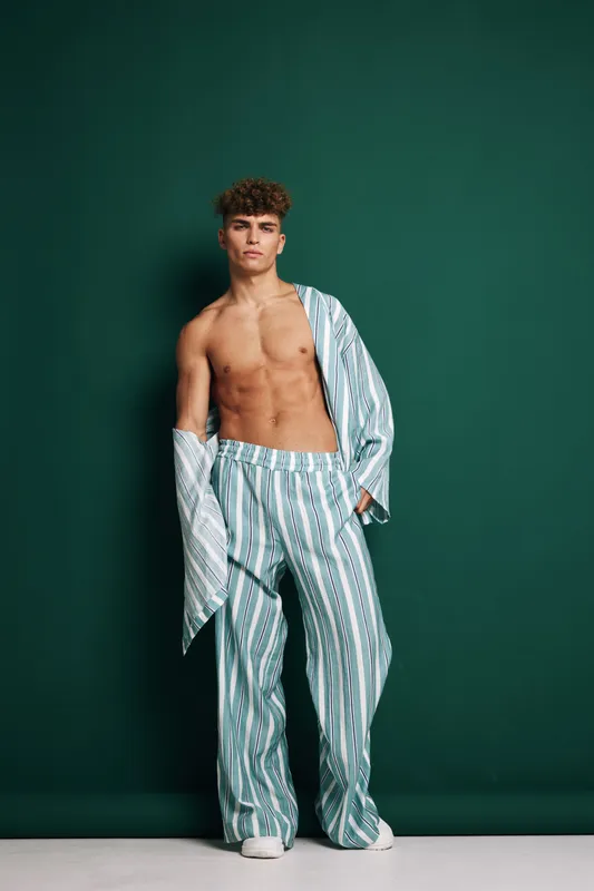 Young male model posing shirtless with striped silky pants and jacket, standing confidently on green background, stylish look with textured curly hair and relaxed expression.