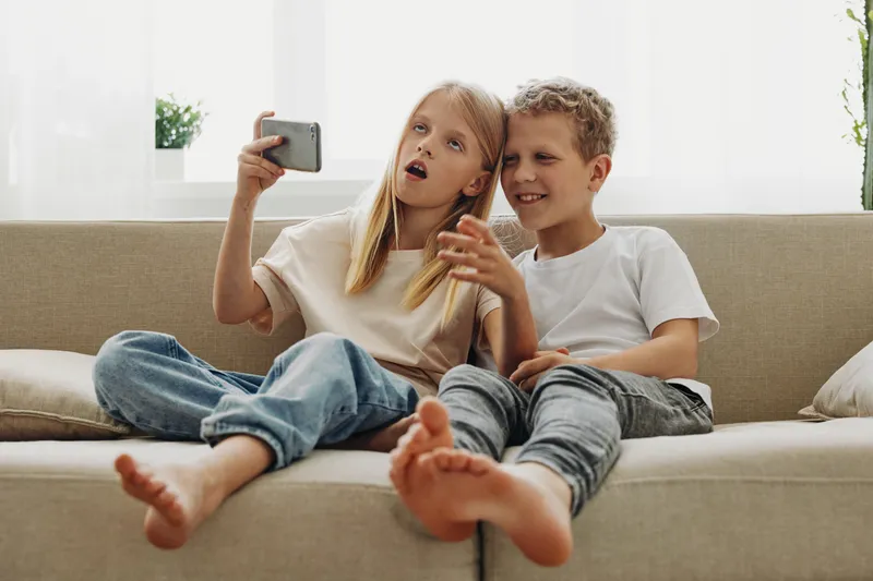 Two happy children sit barefoot on a beige sofa taking a selfie with a smartphone in a bright living room. Concept of childhood fun, friendship, and modern technology use.