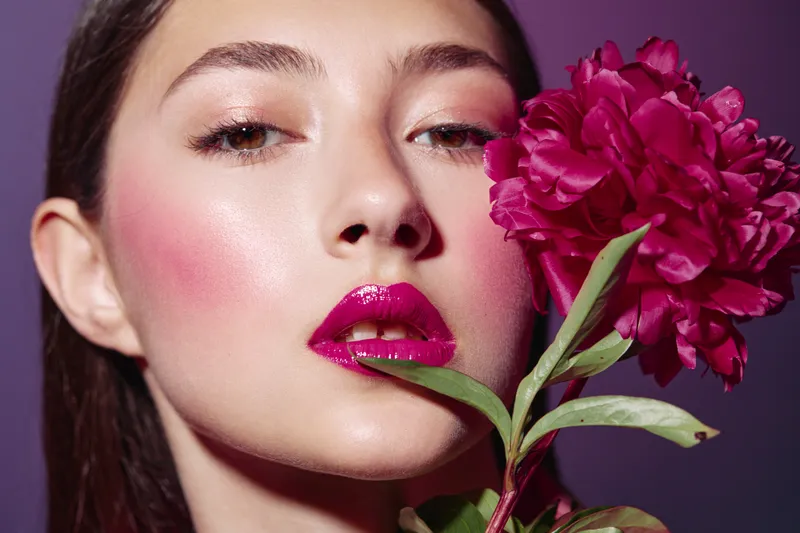 Portrait of woman with glossy pink lips and shiny skin holding vibrant red peony close to face. Beauty concept with creative fashion elements and colorful studio background.