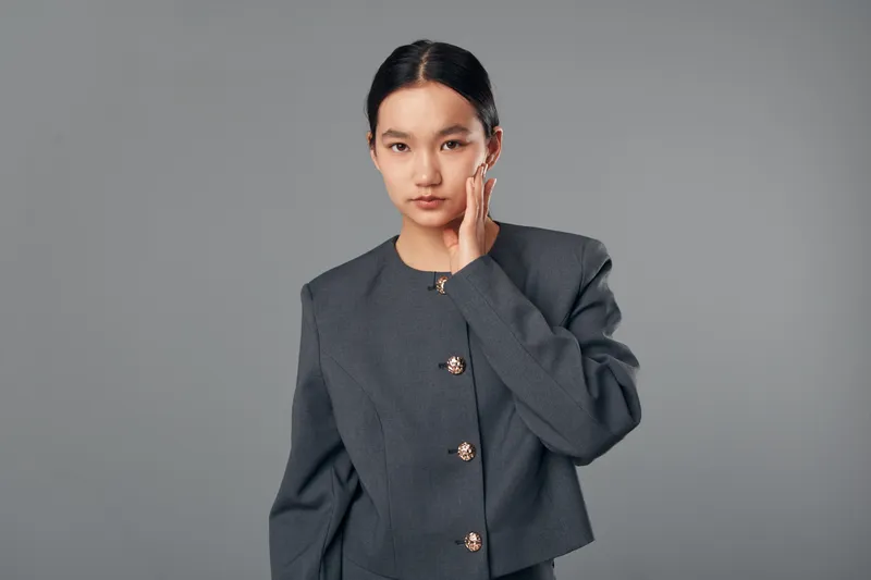 Young woman in gray blazer with minimal makeup and hair tied back posing with hand on face on plain gray background. Female model looking at camera in studio portrait.