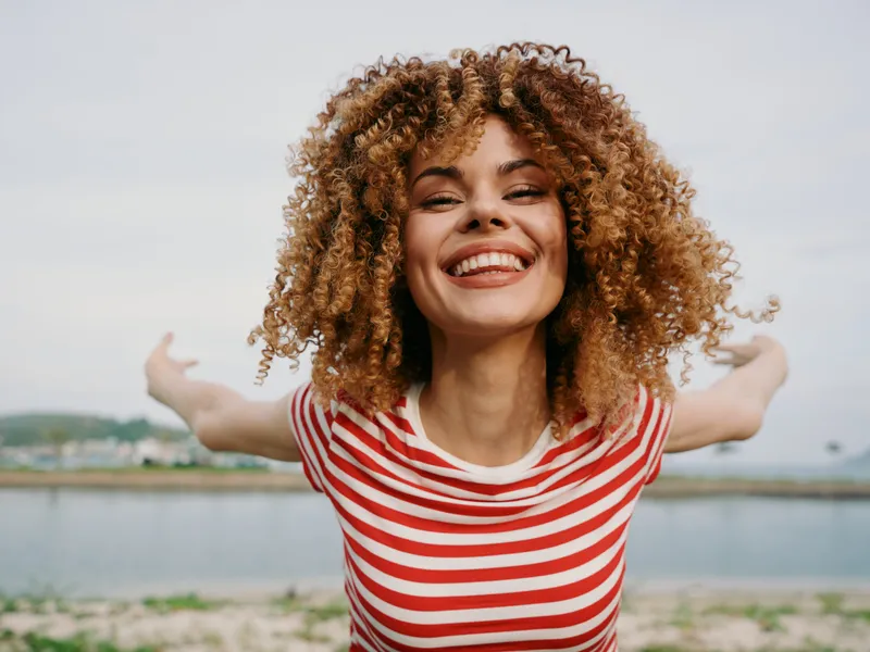 Vibrant portrait of a joyful young woman with curly hair in a red striped shirt, outdoors by a calm waterfront, capturing light, movement, and a carefree summer mood.