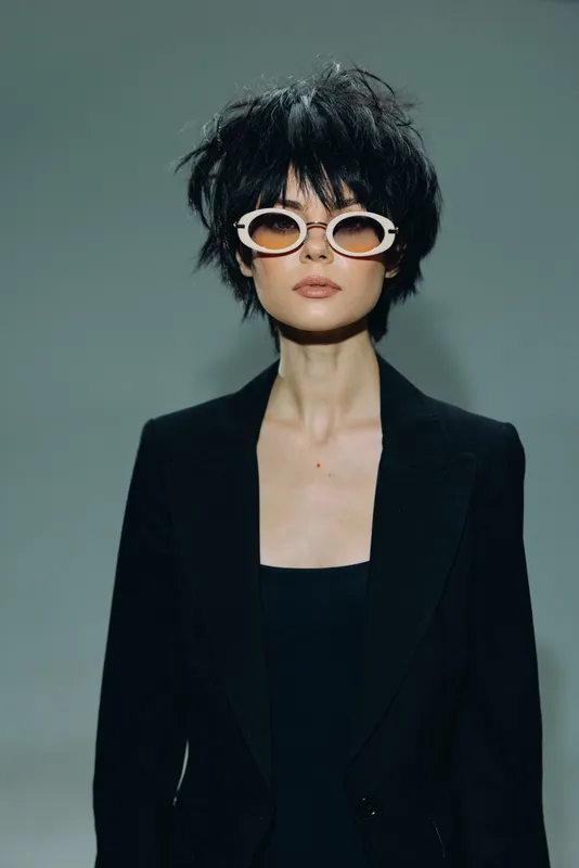 Stylish woman with short black hair wearing trendy sunglasses and a black blazer on a simple gray background.