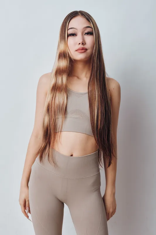 Woman standing and posing with long straight hair wearing beige crop top and matching pants on light background. Female model looking confidently at camera with natural makeup.