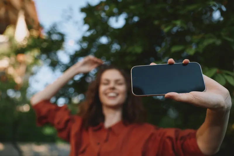 Cheerful woman outdoors, taking a selfie, smiling with eyes closed, sunlit moment, relaxed posture, soft breeze, natural colors, lush greenery in the background