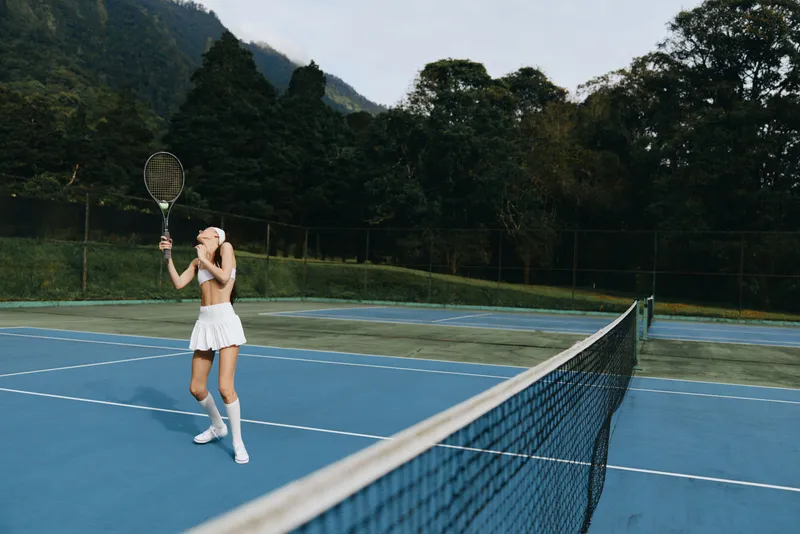 Young female tennis player in white sportswear, playing on an outdoor court surrounded by lush greenery, conveying determination and athleticism