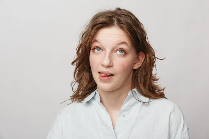 Young woman with curly hair and green eyes makes funny face sticking out tongue. Casual white shirt and neutral background create playful and lighthearted mood in studio portrait.