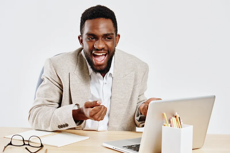 Joyful young man looking at laptop screen with excitement, sitting at desk with notebooks and glasses, expressing happiness and enthusiasm in a modern office environment. People lifestyle concept, pos