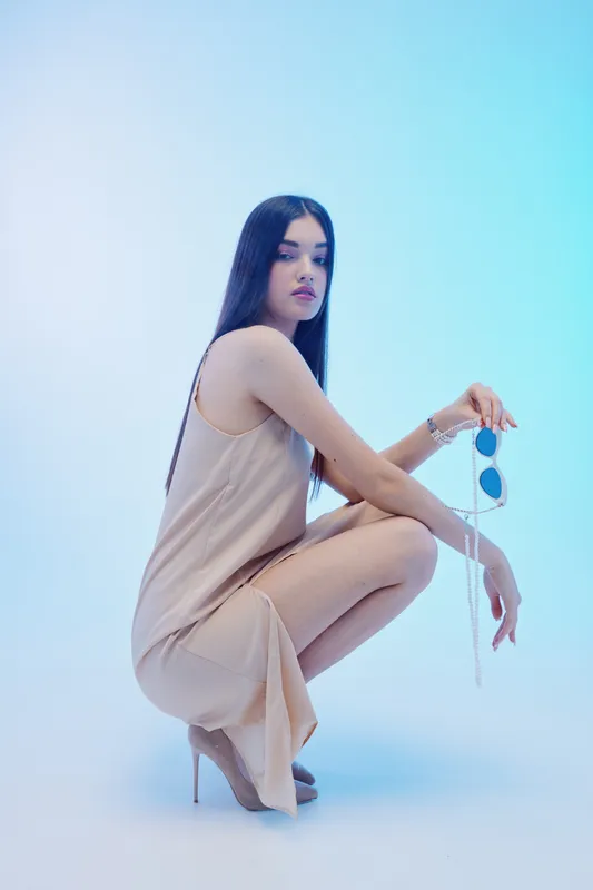 Young woman in a beige dress squats holding sunglasses in a minimalist studio. Her long dark hair and elegant pose highlight beauty, fashion, and style concepts with soft lighting.
