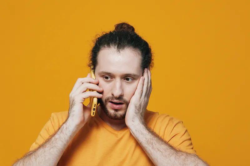 Surprised young man with a shocked expression talking on the phone, wearing an orange t shirt against a vibrant yellow background