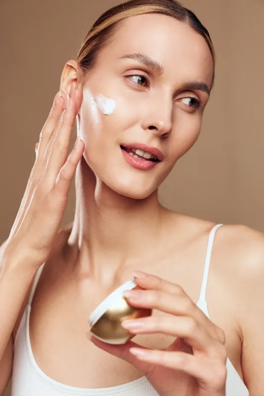 A young woman with a skincare routine applies moisturizer on her cheek in a bright studio, holding a small jar, smiling softly, conveying calm, self care and confidence