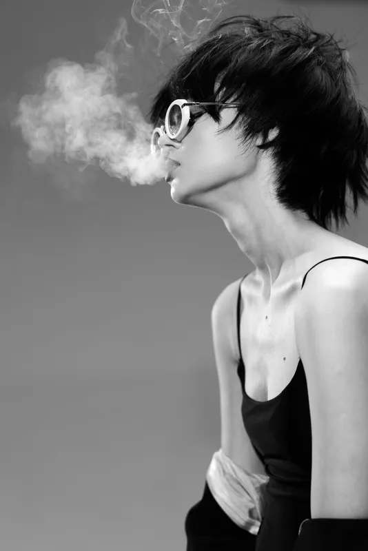 Black and white portrait of a young woman wearing glasses exhaling smoke with a moody atmosphere and stylish look.