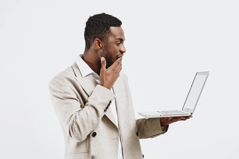 Thoughtful young African American man in casual beige sweater holding laptop computer isolated on plain white background, contemplating or analyzing work, technology device, casual fashion, studio por