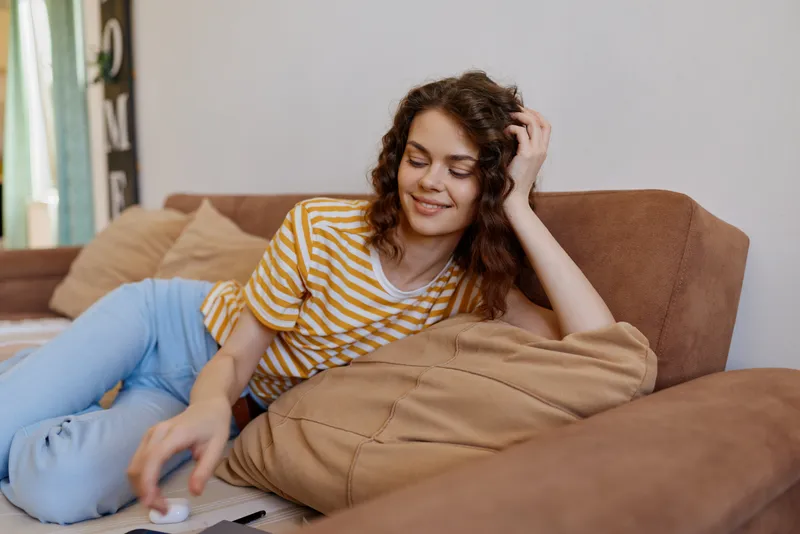 Relaxed woman lounging on a beige sofa at home, wearing striped tee and blue jeans. Casual lifestyle setting emphasizes comfort, relaxation, and everyday moments. Soft natural light highlights a cozy