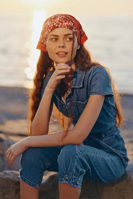 Lifestyle woman in modern boho-western denim street style wearing a red bandana, sitting by the waterfront at sunset in warm film color tones.