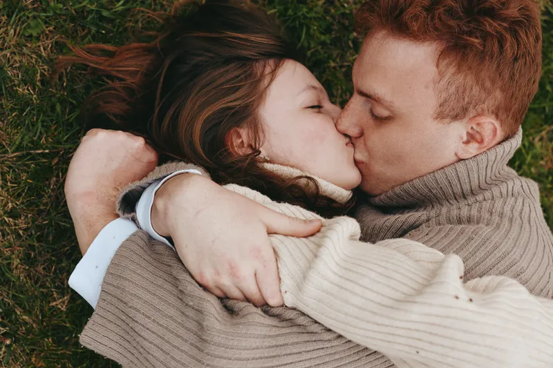 Couple lies on grass outdoors sharing a kiss, wrapped in cozy sweaters, intimate embrace under autumn light, conveying romance, warmth and affection in a candid lifestyle moment.